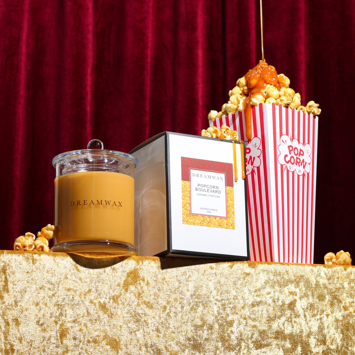 A Dreamwax candle in a clear glass jar with a caramel coloured wax fill and gold label, styled on a gold velvet surface with popcorn and a red curtain backdrop.